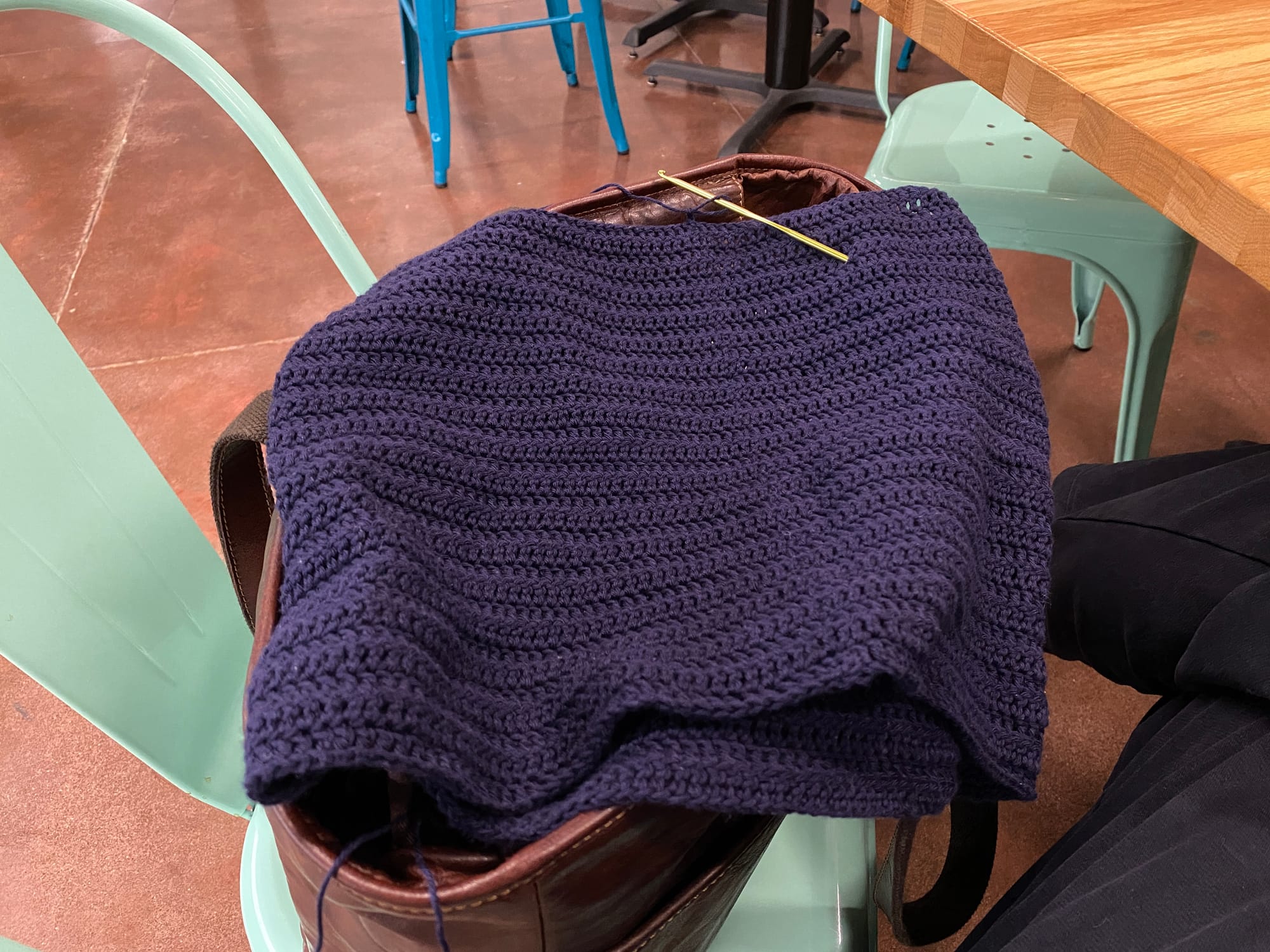 Navy blue crochet worked in the round and about 12 inches in height laid flat over an open brown leather tote bag.  The bag is sitting on a mint green metal chair.