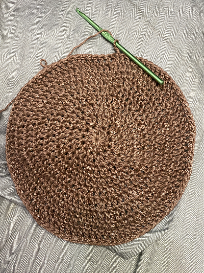 double crochet circle made using brown yarn with a green 4.5mm hook in frame, both resting on light grey blue bed sheets.